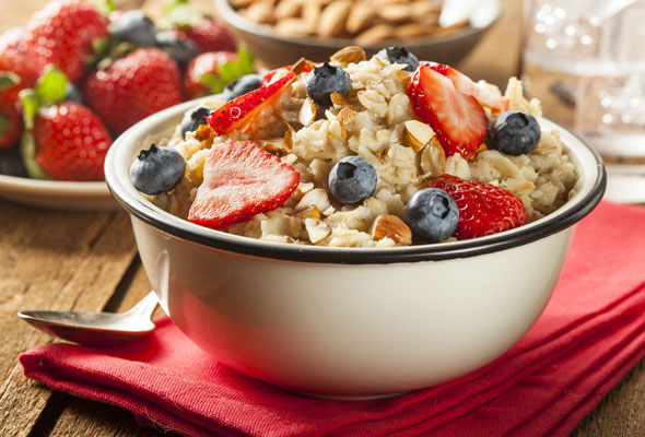 A bowl of oatmeal topped with sliced strawberries, blueberries, and chopped nuts sits on a red napkin with a spoon nearby. Fresh strawberries and a bowl of almonds are in the background.