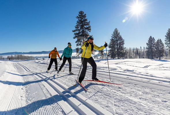 Three people are cross-country skiing on groomed snowy trails under a bright blue sky with the sun shining. They are wearing colorful winter clothing and are surrounded by trees and distant mountains.