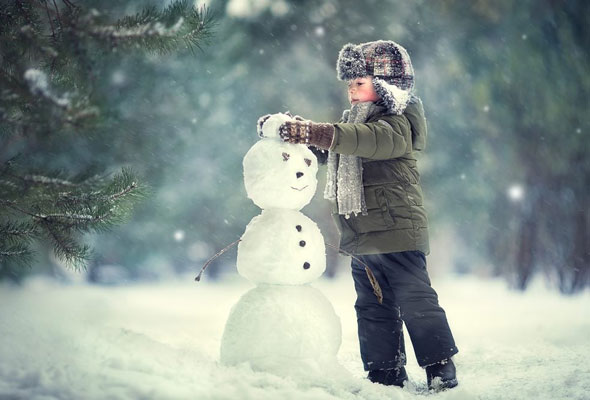 A child wearing a winter coat, scarf, and hat stands in the snow, placing arms on a snowman with a smiling face and stick arms. Snow is gently falling, and trees are visible in the background.