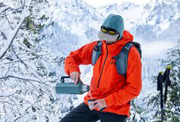 A person in a bright orange jacket and gray hat pours a drink from a thermos while standing in a snowy mountain landscape, surrounded by snow-covered trees. Ski poles are visible in the background.