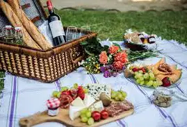A picnic setup on a blanket with a basket, wine, baguettes, glasses, flowers, cheese, fruit, jam, and charcuterie, arranged on plates and boards on grass.