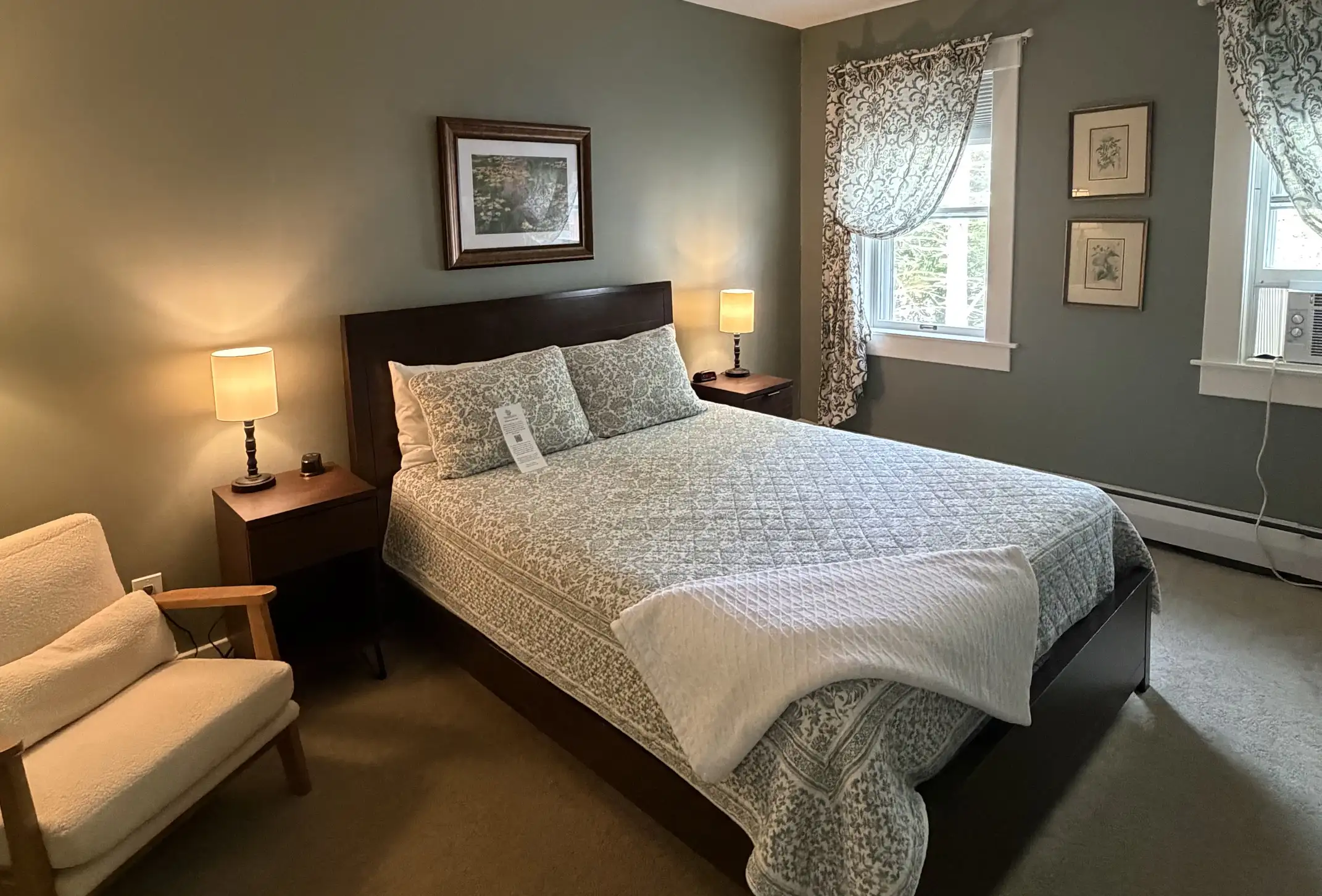 A cozy bedroom with a bed, two bedside tables with lamps, an armchair, framed art on sage green walls, and patterned curtains over two windows. A folded white blanket rests at the foot of the bed.