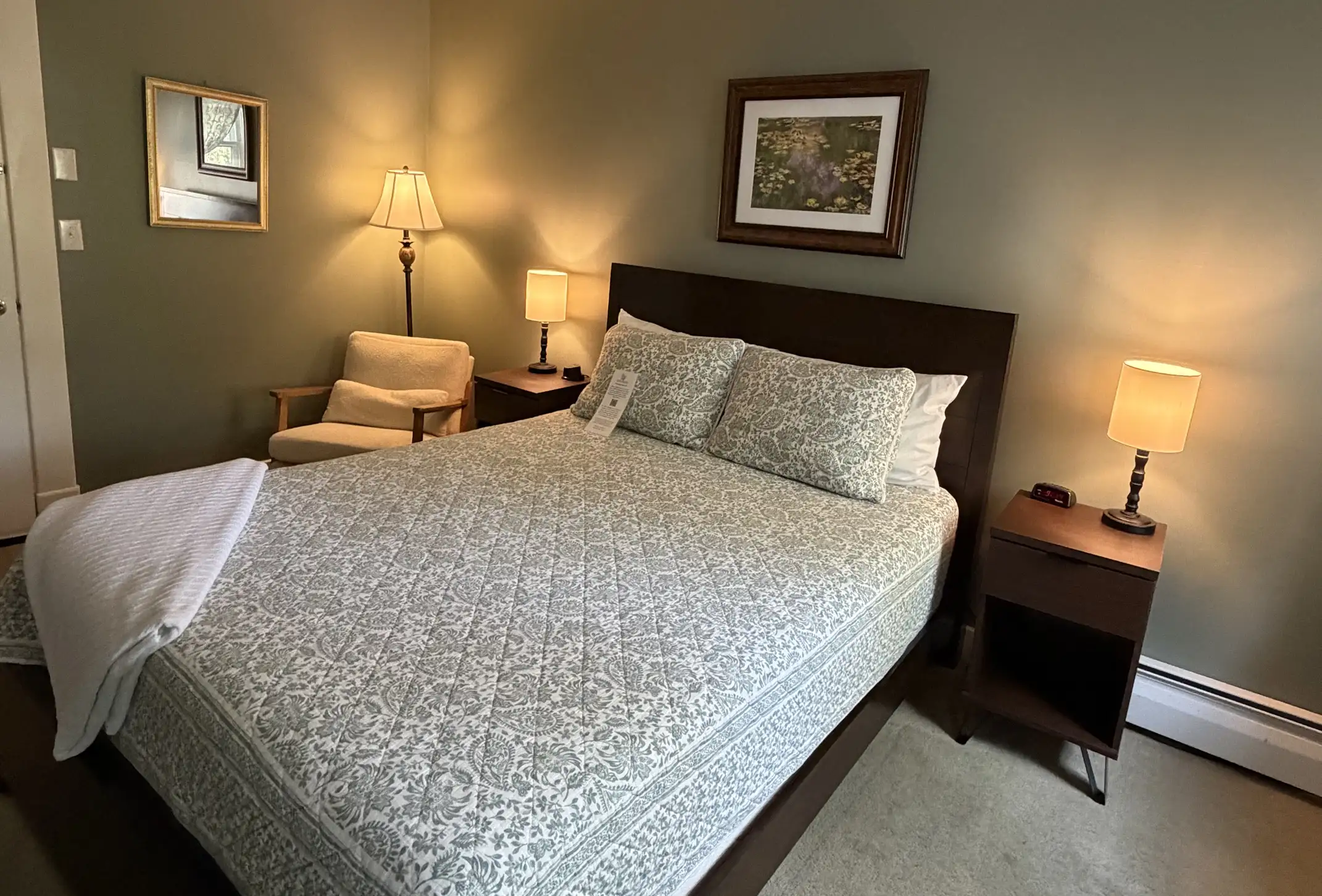 A neatly made bed with patterned bedding in a cozy bedroom, featuring two nightstands with lamps, a framed picture above the bed, an armchair, a standing lamp, and soft green walls.