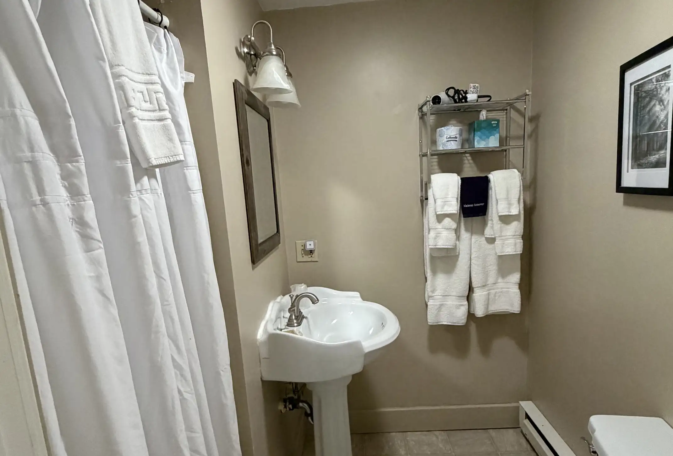A small bathroom with a white sink, mirror, towel rack holding folded towels and toiletries, a shower curtain, beige walls, and a framed black and white photo on the wall.